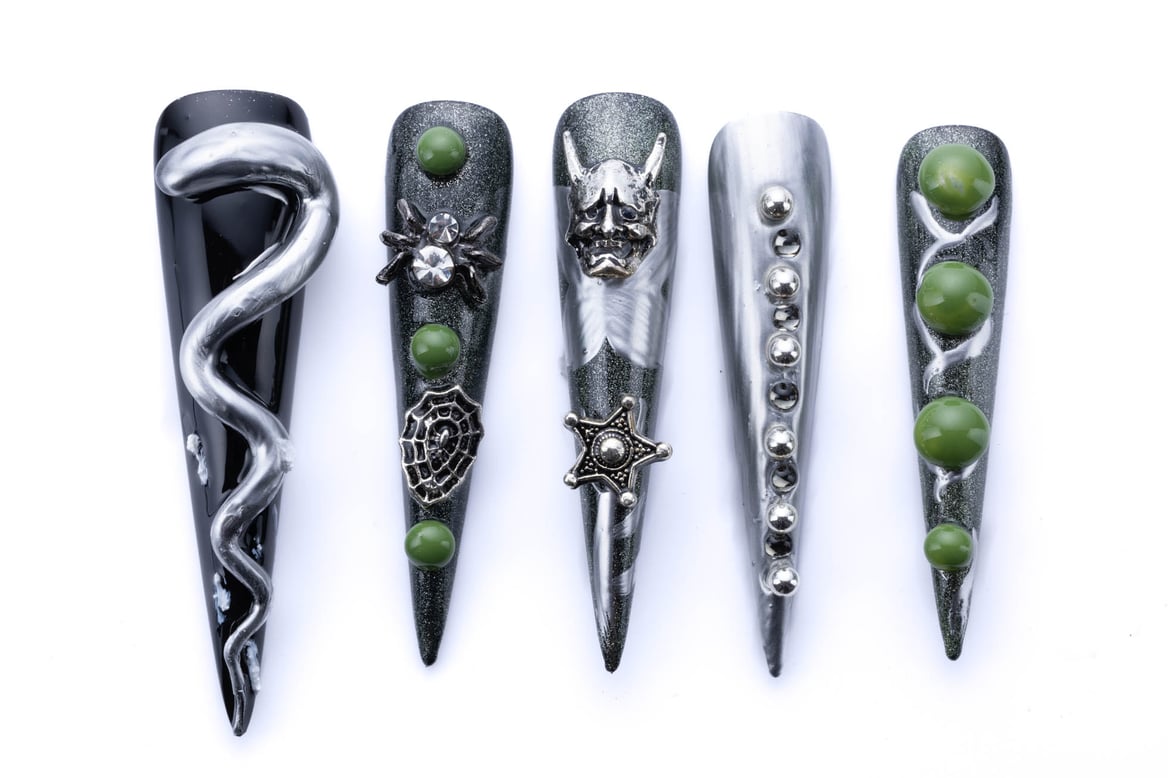 Long stiletto press-on nails in black and green with 3D snake, spider, oni mask, studs, and jewel accents.